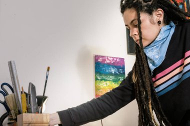 Atölyesinde sanat projesi üzerinde çalışmak için atölyesinde aletlerini tutan, gündelik kıyafetleri ve rastaları olan Latin kökenli genç, beyaz bir sanatçı.