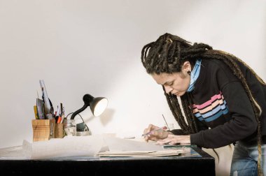 Stüdyoda elinde bir kesici, kağıt ve ahşap bir masa üzerinde çalışma aletleri ile kuaför keserek çalışan oldukça beyaz Latin asıllı genç bir sanat yönetmeni kız..