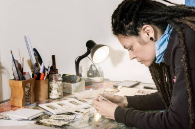 Güzel beyaz bir sanat yönetmeni. Atölyesinde karton ve köpükle çalışıyor. Çalışma masasına yaslanıyor. Masasında kağıtlar ve aletler ve üzerinde parlayan bir lambayla..