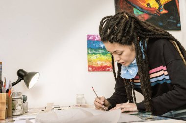 Güzel beyaz kız sanat yönetmeni, İspanyol asıllı, rastalı ve iyi stili olan, atölyesindeki çalışma masasında boya fırçası boyası köpüğüyle çalışırken konuşan..