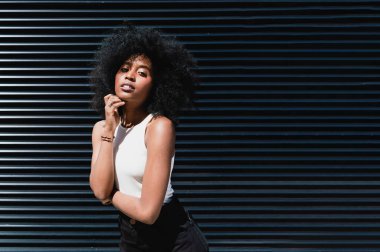 Afro saçlı, siyah-beyaz giyinmiş, metal bir duvarın önünde duran, kameraya bakıp sağdaki boşluğu kopyalayan Latin Amerikalı güzel bir kızın portresi..