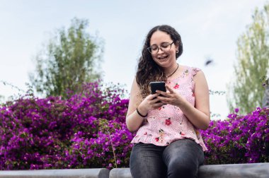 Güzel, genç Latin bir kızın portresi, bukleli ve gözlüklü, güzel mor bir çiçek bahçesinin yanında oturan, telefon, teknoloji ve iletişim kavramını kullanan..