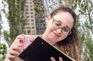 Beyaz Latin kökenli, gözlüklü ve kıvırcık saçlı genç bir kız evinin önündeki parkta oturuyor ve okul defteriyle ders çalışıyor. okul ve yaşam tarzı konsepti.