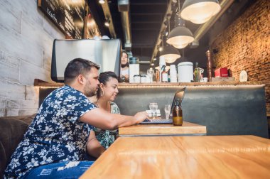 Latin kökenli genç bir erkek ve kadın çift, bir kanepede oturuyor, bir restoranda ahşap bir masanın üzerinde bilgisayar kullanıyorlar, masada alkol jeli spesiyali var..