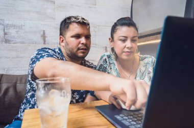 Latin kökenli genç bir çift. İnternette bilgisayar kullanarak çalışıyorlar. Elleriyle ekranı işaret ediyorlar ve kahve dükkanındaki masada içki içiyorlar..