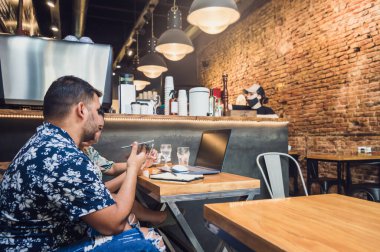 Genç erkek ve kadın çift telefonlarıyla bir restoranda çalışan Latin kökenli serbest işçiler ve ahşap bir masada bilgisayar, internette iş yapan genç işçiler..