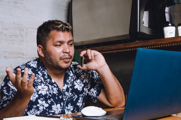 Genç esmer Latin bir adam, kafası karışmış bir halde telefonla konuşuyor ve bilgisayarını kullanıyor, kafeteryada oturuyor, internette iş yapıyor..