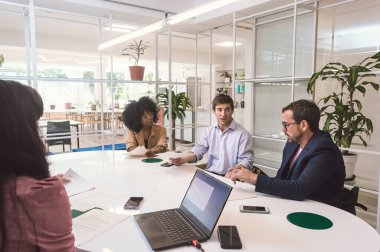 young latin man discussing a contract with the lawyer meeting with other people inside an office of the insurance company, with documents, computer and telephones on the meeting table.