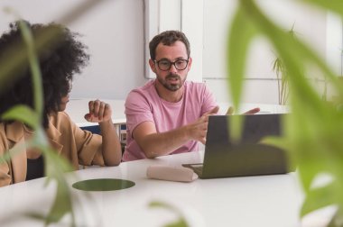 Hispanic Latino adult man, with a beard and glasses, sitting with a computer giving explanations talking to a girl with an Afro, in a business meeting in a coworking.