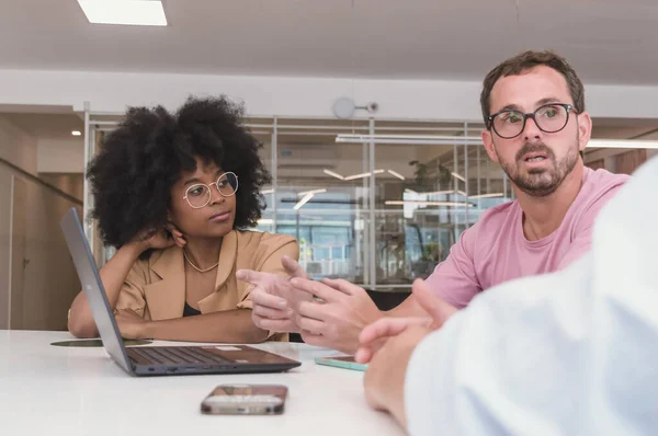 Young Hispanic Latino Afro girl watching a Caucasian Latino adult man with a beard and glasses who is talking and showing his computer, in a business meeting in a coworking, he is giving explanations