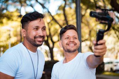 two young, attractive, Hispanic Latin boys with beards and short hair, dressed in white flannel, one of them is holding a stabilizer with a phone in his hand while taking a picture of themselves. clipart
