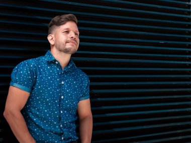 Genç, yakışıklı, sakallı ve kısa saçlı, kısa kollu mavi gömlekli Latin çocuk, profil olarak poz veriyor mavi ve siyah çizgileri olan metal bir duvara yaslanıyor..