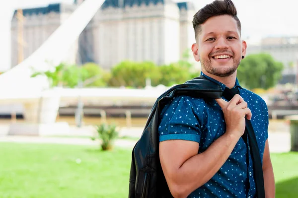 young, attractive, Hispanic Latin boy, with beard and short hair, dressed in a blue short sleeve shirt, holding a black jacket over his shoulder, posing half sideways in a plaza in Argentina.