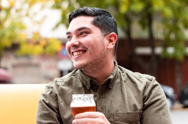 Sakallı ve elinde bir bardak bira olan genç bir latin adam, barın dışındaki sarı bir kanepede oturuyor, gülümsüyor ve iyi vakit geçiriyor. Latin Amerika barı kavramı.