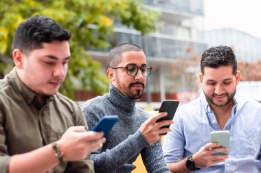 Bir grup Latin arkadaş, telefonlarına bakıyorlar, bir sonbahar günü bir barın dışındaki sarı koltukta oturuyorlar, teknoloji ve yaşam tarzı konsepti..