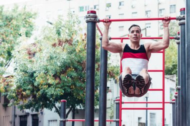 Genç atletik atlet, beyaz tenli, aynı anda barfiks çekiyor ve mekik çekiyor, arka planda gökyüzü, spor salonunda, açık havada.