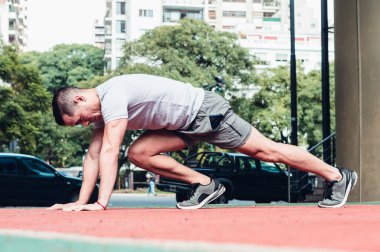 Beyaz tenli ve gri giyimli genç Latin atlet yan yatmış, kardiyak pozisyonunda egzersiz yapıyor, dizlerini bir fitness plazasında göğsüne getiriyor..