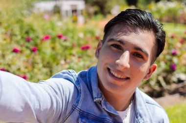 Kafkas kökenli Latin kökenli genç bir adamın halk parkında arka planda güllerle selfie çektiği yakın plan portresi.,.