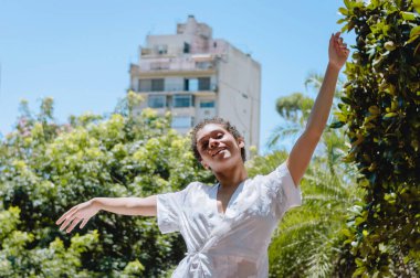 Parkta kısa saçlı ve beyaz elbiseli genç Arjantinli kadın kameraya bakıp gülümsüyor. Kollarını açıp özgürlüğü, kadın dahil olmayı ve yaşam tarzı kavramını hissediyor..