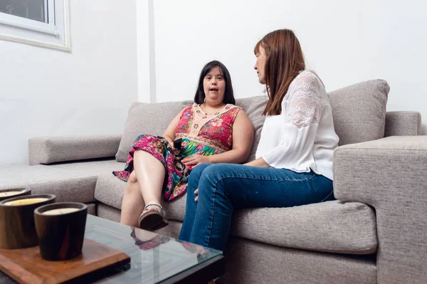 disabled caucasian adult woman at home sitting on the living room sofa talking to her mom, domestic life between mother and daughter with down syndrome.