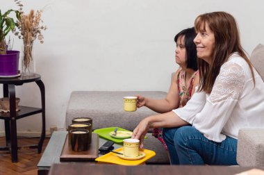 Concentrated young latin caucasian woman with down syndrome watching television with her mom sitting in the living room and holding some cups of coffee. clipart