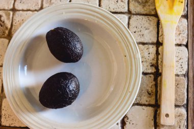 İki avokado, porselen bir tabakta, mutfak tezgahındaki ahşap bir spatulayla yan yana..