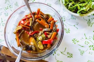 Masanın üzerinde pişmiş sebzelerle dolu cam kase, yanında da taze roka..