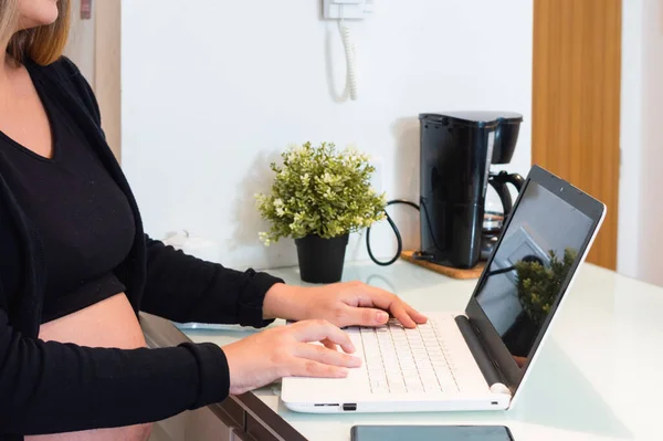 Hamile beyaz bir kadının mutfak tezgahında dizüstü bilgisayarda yazı yazması, online iş yapması, e-postaları kontrol etmesi ve online alışveriş yapması..