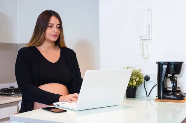 妊娠中の若い白人女性は台所のカウンターで彼女のラップトップでオムツをオンラインで購入し 割引やオファーのためにネットをサーフィンします 顧客やライフスタイルのコンセプト — ストック写真