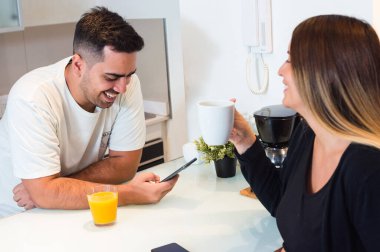 Kafkasyalı genç adam tezgaha yaslanmış telefonla gülüyor ve mutfakta eşiyle konuşuyor, heteroseksüel çift evde gülüyor ve birlikte eğleniyor..