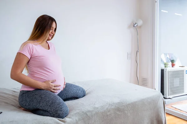 妊娠中の白人の若い女性が彼女のベッドの上に座って目を閉じて腹に触れるだけで出産の準備を瞑想産前のコンセプトコピースペース — ストック写真