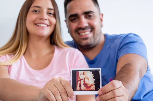 怀孕的高加索男人和女人笑着看着摄像机模糊了背景 显示了打印的超声波照片 年轻的异性恋夫妇为他们的新儿子感到高兴 — 图库照片