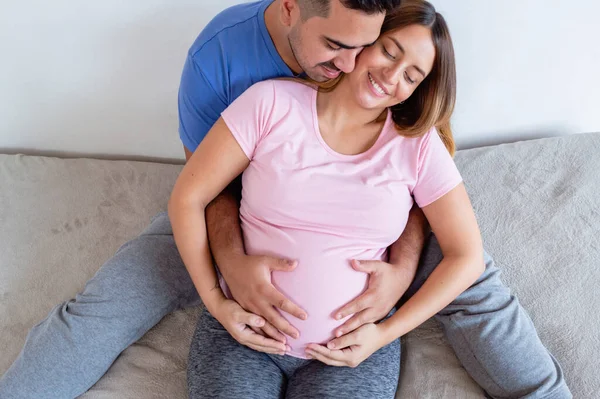 トップ表示妊娠中の白人カップル抱き合って目を閉じてベッドに座って壁にもたれかかって幸せな笑顔で腹に触れて — ストック写真