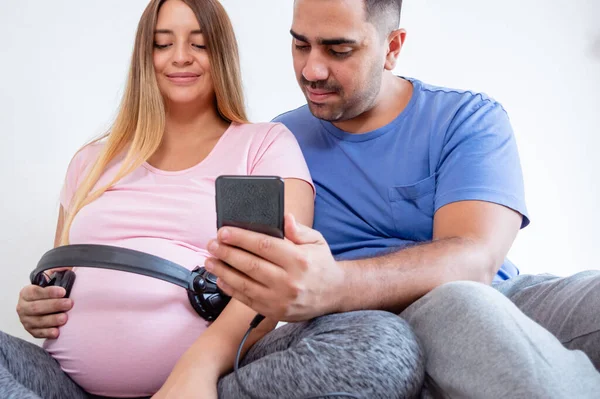 妊娠中の若いベネズエラの白人カップルは一緒に彼の腹にヘッドフォンで彼らの赤ちゃんに音楽を教えてベッドの上に座って 音楽を再生するために携帯電話を使用して 妊産婦の概念 — ストック写真