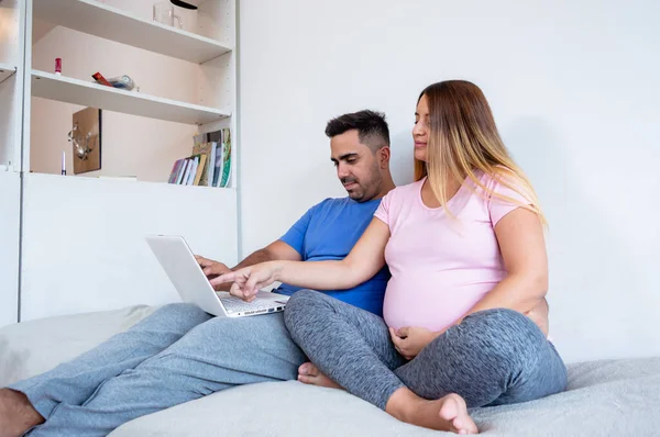 妊娠中の白人の若い女性ピンクとグレーのパジャマ夫とベッドに座って青いパジャマを着た髭の男彼らはラップトップを使ってビデオを見てネットサーフィンをしています — ストック写真