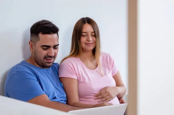 サイドビュー若いベネズエラ人男性と一緒に座っている彼の妊娠中の妻とパジャマで部屋に座って ベッドの上に座っているビデオや映画をラップトップで見て インターネットサーフィン — ストック写真
