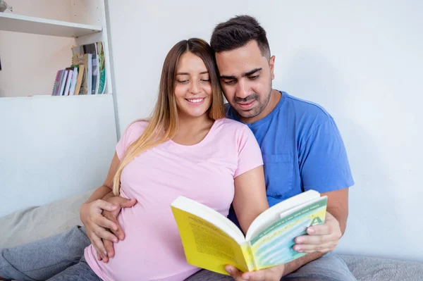 前景色 穿着睡衣的怀孕夫妇的肖像一起坐在床上看书 身穿蓝色衣服的男子与妻子触摸腹部 躺在床上看书 — 图库照片