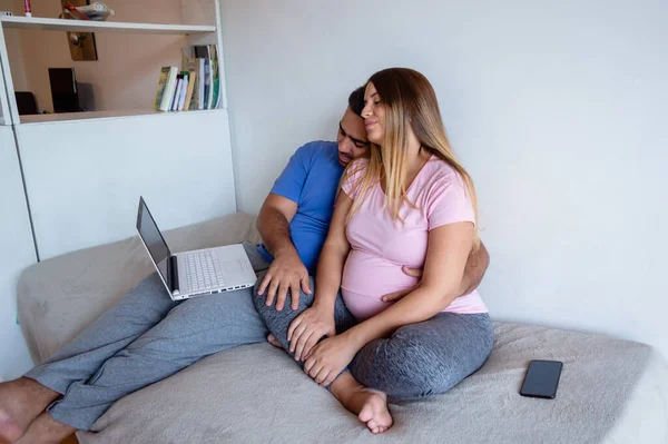 怀孕的白种人夫妇坐在床上 使用笔记本电脑 上网观看系列节目 在家里享受一天的假期 — 图库照片