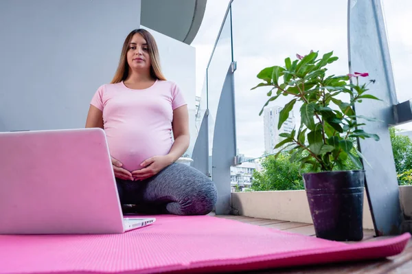 妊娠中の白人の若い女性がピンクのヨガマットに座っています自宅のバルコニーでラップトップでオンラインヨガクラス コピースペースと概念オンラインクラス — ストック写真