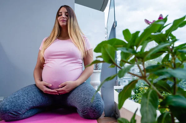 若い妊娠中の白人女性目を閉じた瞑想マットの上で彼女の腹に触れる彼女の家の中の前景に植物 コピースペース — ストック写真