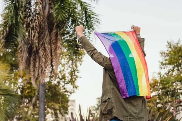 rear view unrecognizable man outdoors holding gay pride flag on his back with his hands, copy space.