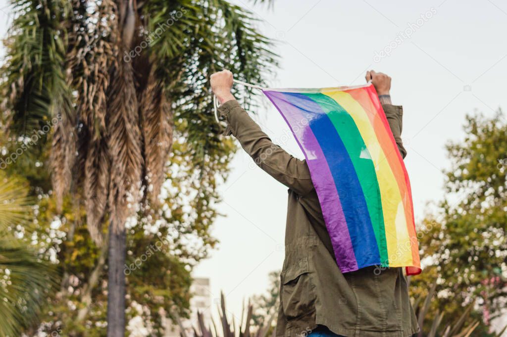 rear view unrecognizable man outdoors holding gay pride flag on his back with his hands, copy space.