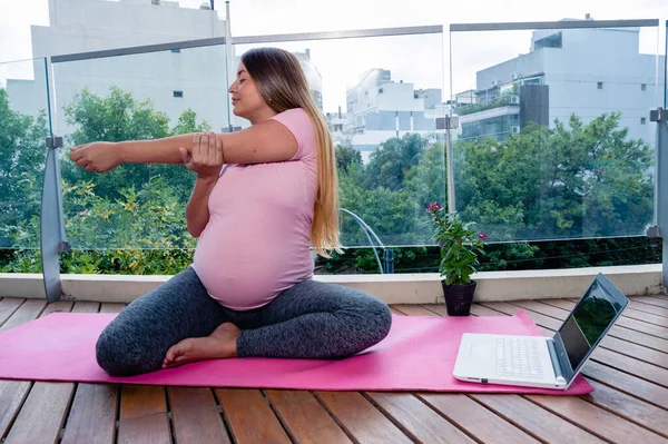 妊娠中の若い女性はマットの上に彼女のアパートのバルコニーに座って彼女のラップトップでオンラインヨガクラスで腕の伸びをやって — ストック写真