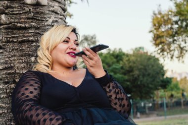 Beyaz Arjantinli genç kadın portresi. Açık havada oturmuş gülümsüyor. Telefonla ses notu gönderiyor..