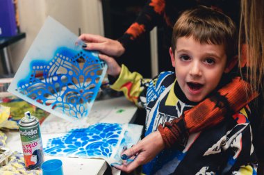 Mutlu çocuk annesiyle oynuyor, evde mavi spreyli kağıtları boyuyor, ağzı açık kameraya bakıyor..