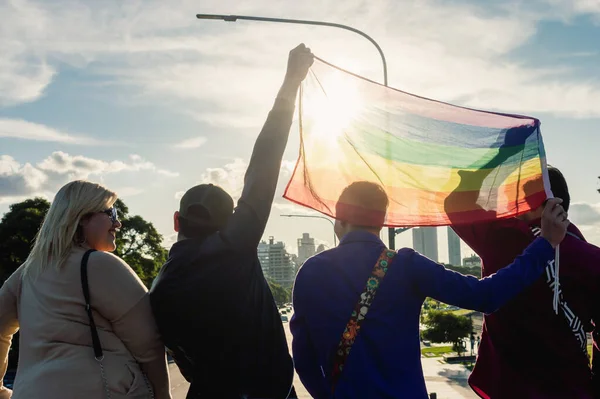 Bir grup erkek ve kadın arkadaşın arka planı, ellerinde gay bir gurur bayrağı tutuyorlar, bayrağın arasından güneş parlıyor, gökyüzü ve şehir panoramik bir manzara ile.