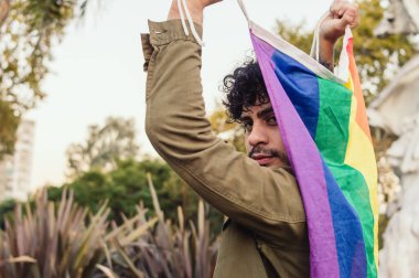 Beyaz bir adamın arka görüntüsü, elinde gay gururu bayrağı, kameraya bakıyor..