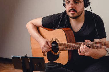 Beyaz tenli, gözlüklü genç bir adam yerde oturmuş telefonda gitar dersi izliyor, akustik bir gitarla pratik yapıyor ve çalışıyor..