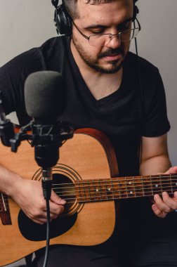 Kafkas asıllı, gözlüklü, kulaklıklı, siyah elbiseli, müzik stüdyosunda oturan, akustik gitar çalan ve daha yoğun bir mikrofon kullanarak şarkı söyleyen Latin bir adam. portre resmi.