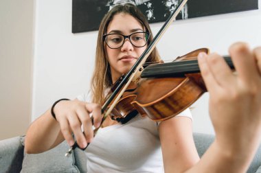 Beyaz giyimli, keman çalan, evdeki oturma odasındaki kanepede oturan, gözlüklü, beyaz tenli, genç, beyaz tenli, beyaz tenli kadın..